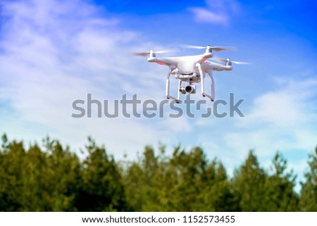 White is flying high in the air, taking photos and recording footage from above. Flying drone with four motors and propellers, camera and red warning lights on clear blue sky background. 