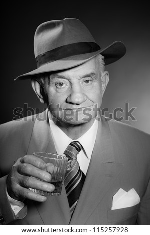 Senior glamour vintage man wearing suit and tie and hat. Black and white studio shot. Gangster look. Drinking glass of whisky. Isolated.