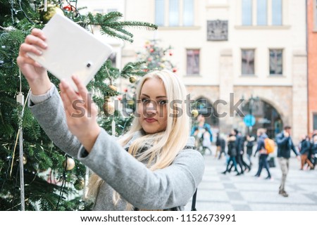 A beautiful young blonde woman or girl doing selfie or photographing next to a Christmas tree during Christmas holidays at the Old Town Square in Prague, Czech Republic.