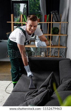 smiling young cleaning company worker cleaning sofa with vacuum cleaner and showing thumb up