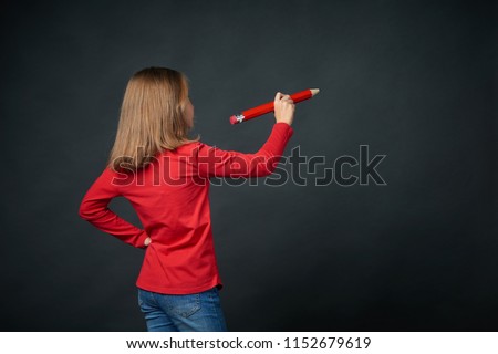 Back view of a school age girl holding big pencil and drawing or writing on blank copy space for text