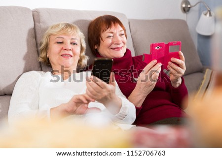 Smiling pensioners females on sofa holding mobile phones in hall