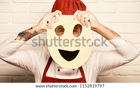 Cooking process concept. Chef makes pizza. Cook with hidden face in burgundy uniform plays with dough. Man with beard holds rolled dough with cut smiley face on white brick wall background.