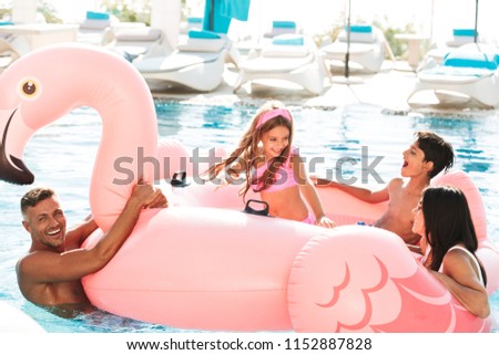 Cheerful young family having fun together at the swimming pool outdoors in summer, swimming with inflatable flamingo