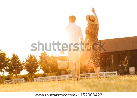 Back view picture of young loving couple with basket of drinks and food going to have picnic outdoors.