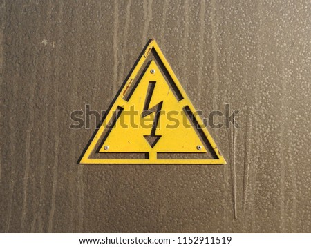 
A sign of high voltage in the form of a metallic relief attached to a brown steel surface.					