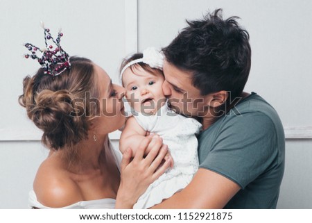 Parents hold the child in their arms between each other and kiss the baby. Man, woman in crown and daughter on white background