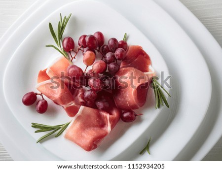 Prosciutto served with rosemary and grape on a white plate . Top view.