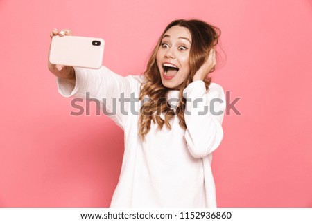 Portrait of an excited young woman taking selfie with mobile phone isolated over pink background
