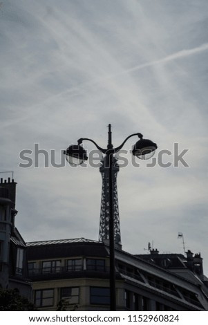 Parisian funny cityscape of top of Eiffel Tower silhouette mixed with a street lamp, making an optical illusion of a giant bug, or insect, with big eyes and a long neck, under grey cloudy sky, Paris.