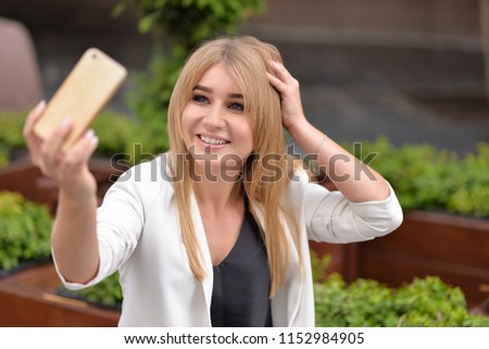 A girl with blond hair in a white jacket and black blouse smiles and makes selfie. Focus on the face.
