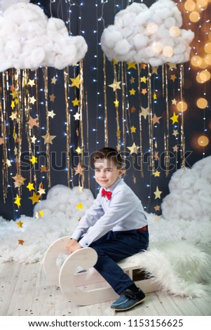 Magic christmas card with a boy in a studio with a gold stars garland, fluffy clouds and lights. Happy boy with red bow tie sits on a white wooden sleigh