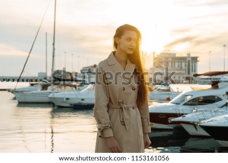 a young thoughtful girl with long hair, in a beige fashion coat is waiting for her yacht in the seaport