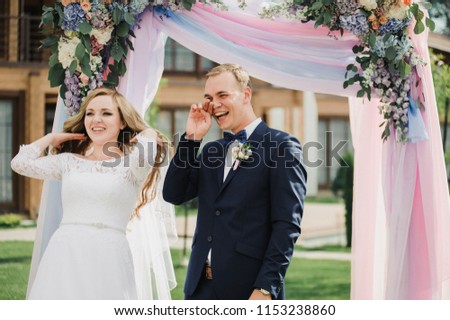 Wedding ceremony in the open air. An arch of real flowers. hydrangea and roses. Pink and blue fabric. The bride and groom are exchanging oaths. Blue suit, bow tie, white fluffy dress and veil.