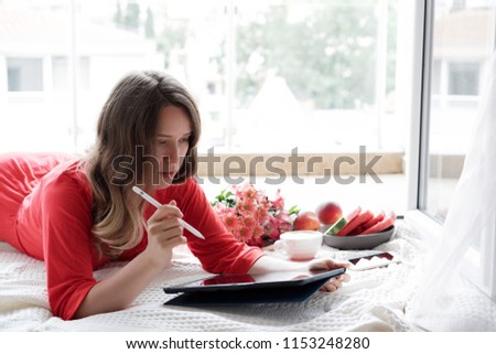 Pretty blonde woman in a red robe working on tablet on the bed and drinks coffee against window background 