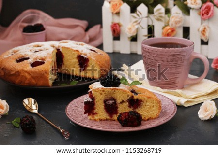 Pie with blackberries is located on a dark background. In the foreground is a piece of cake. In the photo there is a cup and a fresh blackberry.