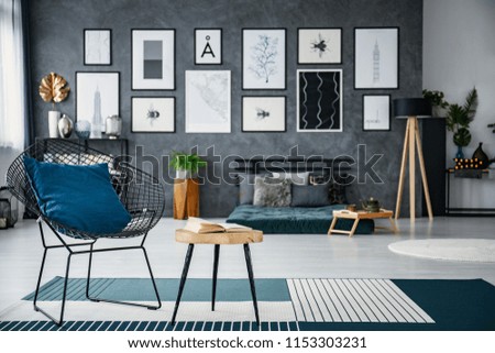 Blue pillow on armchair next to table in living room interior with futon and posters. Real photo with blurred background
