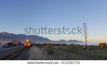 A cargo train departing from train station during twilight hours in Kaikoura, New Zealand. The color of the sky is soft and vibrant. In the background, there are mountain, ocean and a unique tree. Royalty-Free Stock Photo #1153325710