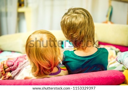 blonde baby girl and dark hair boy watch cartoon on tablet PC back view. Brother and sister with tablet PC at home. Little boy and girl sitting on sofa and watching cartoon on mobile in the evening