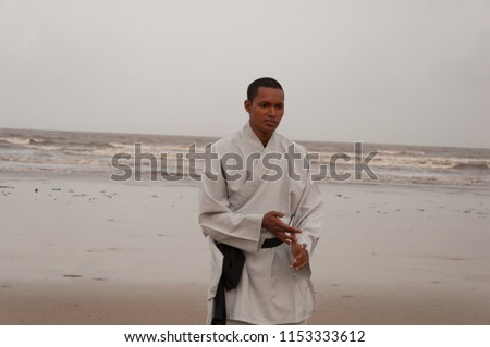Full length body action shot of young Asian male Shaolin Kung Fu instructor training on Juhu beach, Mumbai. Intense concentration shows on his face. Sand in foreground and grey sky in background.