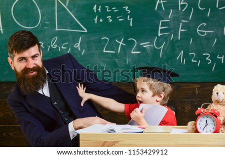 Father with beard, teacher teaches bored son, little boy. Boring studying concept. Teacher and pupil in mortarboard, chalkboard on background. Kid fed up with studying, kicks away teacher.