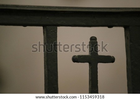 Close up view of a small metallic cross incorporated in a black forged gate. Religious sign located in a french church. Silhouette of a catholic symbol. Blur brown background. Abstract picture.