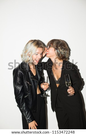 Two beautiful smiling girls in black jackets with glasses of champagne in hands happily gossiping over white background