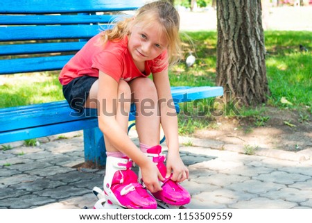 Joyful girl, blond clothes rollers of red color and looks at the camera and smiles, sitting in the park on a blue bench