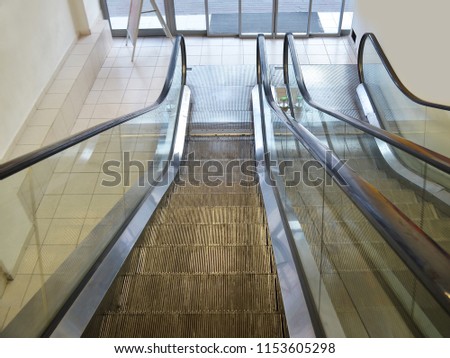 Escalators from the ground floor to the second one inside a supermarket, top-down view. The walls are painted in a light yellow color. Sliding glass doors lead to the street
