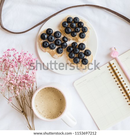 Flatlay items: monthly planner, pen with flamingo, plate with bread and blueberries, cup of coffee, flowers pink color, wicker bag lying on white background