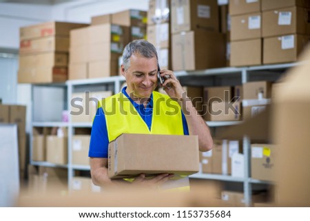 Factory worker. Smiling warehouse workers preparing a shipment in a large warehouse. Worker In Distribution Warehouse. Worker marking shipping list and carrying cardboard box for delivery 