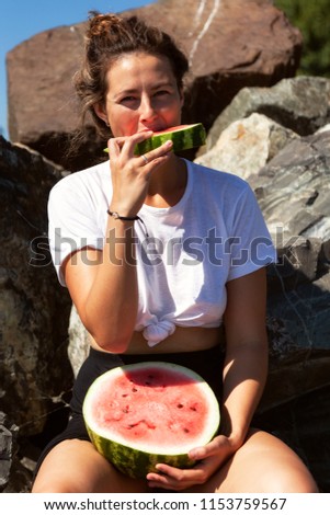 young beautiful woman laughing happily, smiling, holding between the legs a big watermelon and eating, sitting stone rocks on a summer day near the sea
