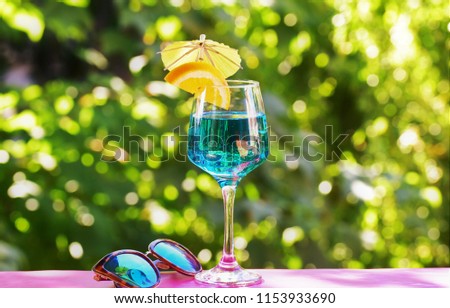 Blue drink in a beautiful wine glass placed in the back porch.With sunglasses in front with green nature.
