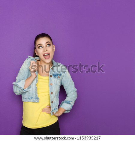 Excited beautiful young woman in jeans jacket is looking away and talking. Waist up studio shot on purple background.