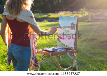 A young woman artist holds a brush and paints a picture on an easel in the rays of the sunset. The painter paints oil paintings in the garden at sunset