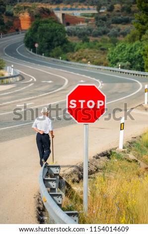 Elderly man walking with a cane past a red stop sign leading to a curving highway. Conceptual metaphors: forever young, never being too old to follow your dreams, defiance, overcoming challenges. etc
