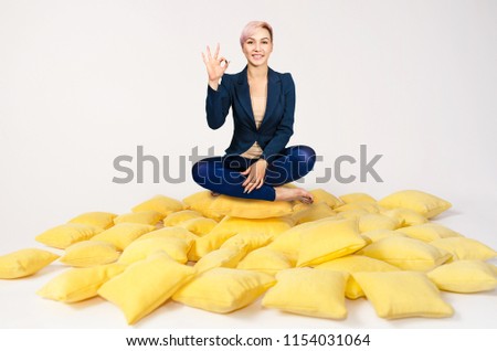Young beautiful girl sits in a lotus pose on a pile of yellow pillows and shows sign ok, on a white background.
