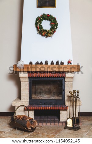 modern fireplace decorated with a Christmas wreath and a Santa Claus statue. Before the fireplace there is a forged drovnitsa and a set of brass tools
