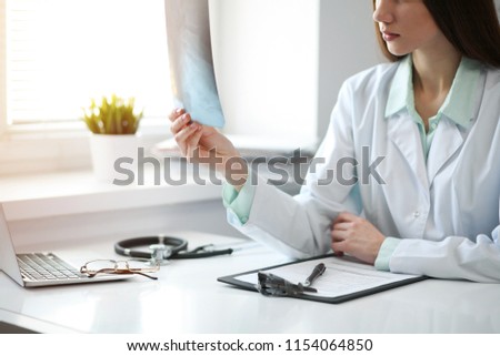 Young female doctor examining x-ray while sitting at the table near the window in hospital office