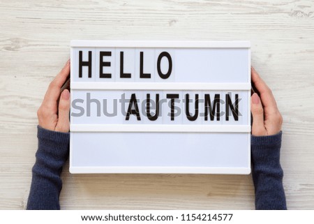 Female hands hold modern board with text 'Hello Autumn' over white wooden surface, top view. Overhead, flat lay, from above.
