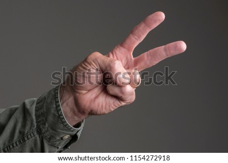 Isolated man's hand, two fingers up in peace sign.  Green canvas shirt buttoned at the cuff.  Dark grey background.