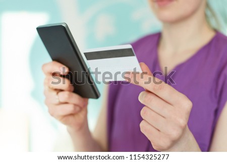 Cropped image of modern smart phone and plastic card in woman`s hands against blue blurred background. Young businesswoman checks her bank account in mobile application. Online payment concept