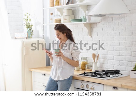 Young woman using smartphone leaning at kitchen table with coffee mug and organizer in a modern home. Smiling woman reading phone message. Brunette happy girl typing a text message Royalty-Free Stock Photo #1154358781