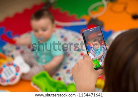 Woman photographing her baby with a smartphone while he plays with toys.