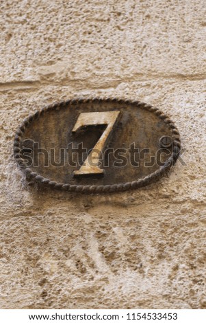 Close up outdoor view of the number seven written in beige on a brown metallic oval plate. Numerical symbol fixed on a wall and indicating the address of a house in a french street. Ancient object.