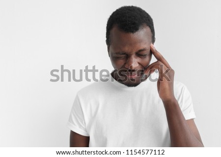 Horizontal portrait of African American man pictured against grey background feeling severe headache he is suffering from, touching his head, bending head and grinning with closed eyes because of pain