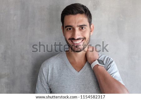 Indoor closeup of young good looking European man standing against gray textured wall, dressed in t-shirt, showing friendly toothy smile,  satisfied and confident 