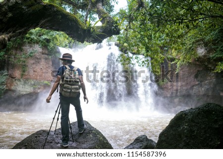Young asian man standing photographing a waterfall, Traveler standing on the rock and photographing waterfall in the rainforest national park at Thailand.