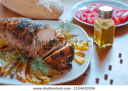 Meat roll with spices in a plate lies on a table. Round the meat roll is baked potatoes. The table is served with fresh tomatoes, olives, black pepper, greens.