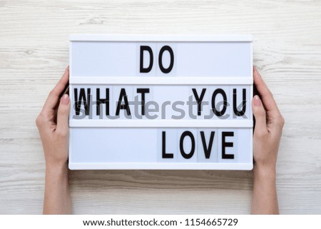 Female hands hold modern board with 'Do what you love' word on lightbox over white wooden background, from above. Closeup.